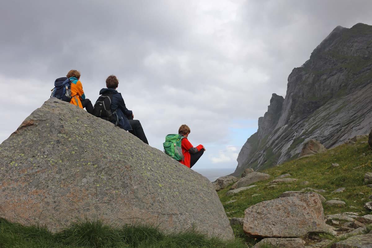 Three hikers resting on a large boulder near Bunes Beach in the Lofoten Islands under cloudy summer skies