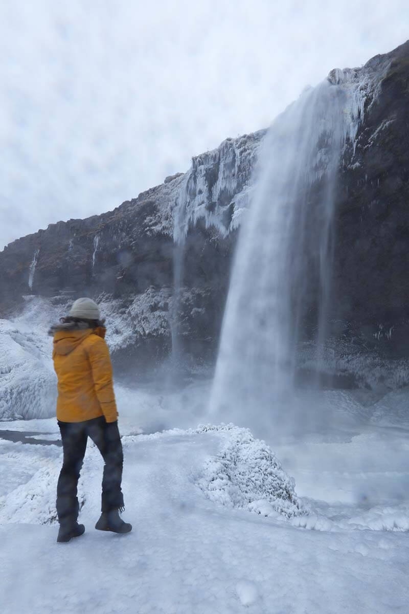 Woman standing at a frozen Seljalandsfoss Waterfall on a rainy cold winter day